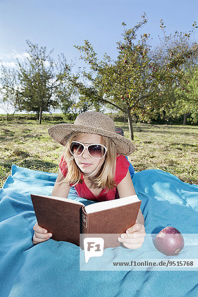 Mädchen  neun Jahre  mit Sonnenhut und Sonnenbrille liest ein Buch auf einer Wiese mit Apfelbäumen
