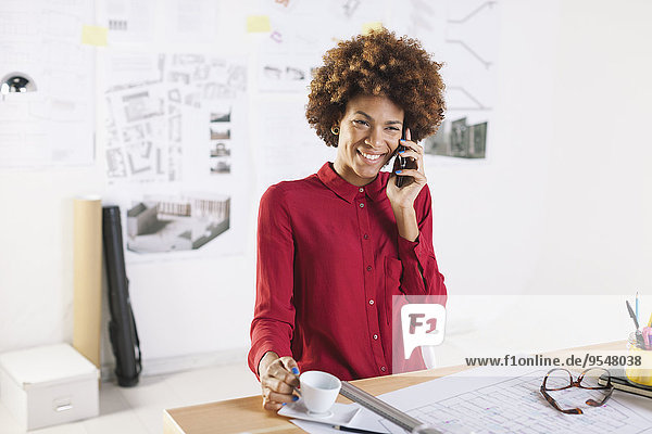 Porträt einer lächelnden jungen Architektin beim Telefonieren mit dem Smartphone in ihrem Büro