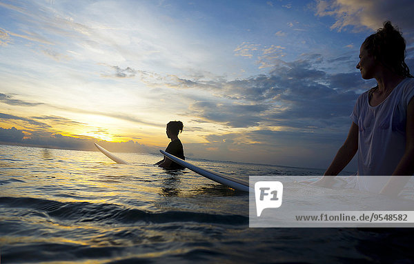 Indonesien  Bali  Canggu  zwei Surferinnen im Wasser  die die Sonne beobachten.