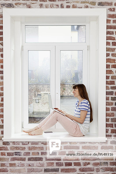 Entspannte Frau sitzt auf der Fensterbank mit Laptop