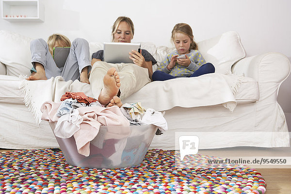 Mutter  Tochter und Sohn auf Couch mit digitalem Tablett und Handy mit Wäschekorb auf dem Boden