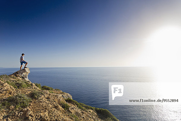 Spanien,  Balearen,  Mallorca,  ein Teenager,  der auf einem Felsen an der Steilküste steht und den Sonnenuntergang beobachtet.