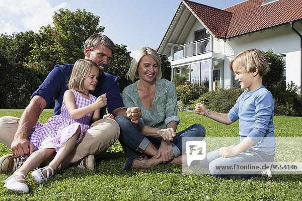 Fröhliche Familie sitzt auf Rasen im Garten und spielt Stein-Papier-Schere
