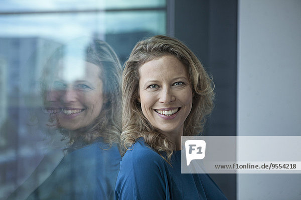 Porträt einer lächelnden reifen Frau  die sich am Fenster in ihrer Wohnung lehnt.