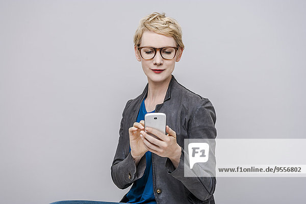 Porträt einer lächelnden blonden Frau mit Smartphone vor grauem Hintergrund