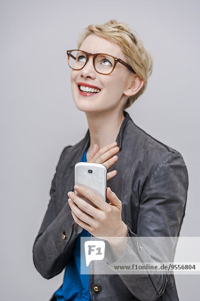 Porträt einer lächelnden blonden Frau mit ihrem Smartphone vor grauem Hintergrund