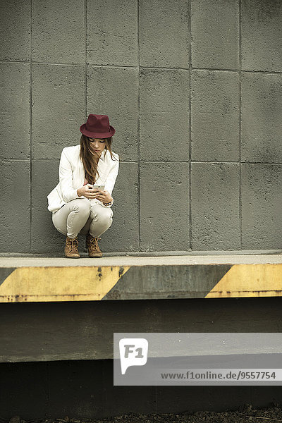 Junge Frau mit rotem Hut  die auf einer Rampe hockt und auf ihr Smartphone schaut.