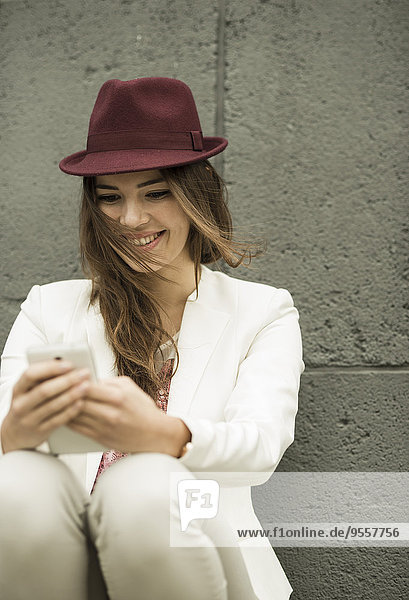 Porträt einer lächelnden jungen Frau  die ihr Smartphone vor der grauen Wand betrachtet.