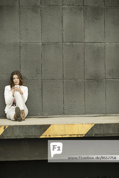 Junge Frau sitzt auf einer Rampe vor grauer Wand mit ihrem Smartphone