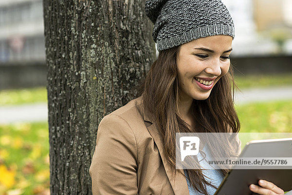 Porträt einer lächelnden jungen Frau  die sich mit einem digitalen Tablett an einen Baum lehnt.