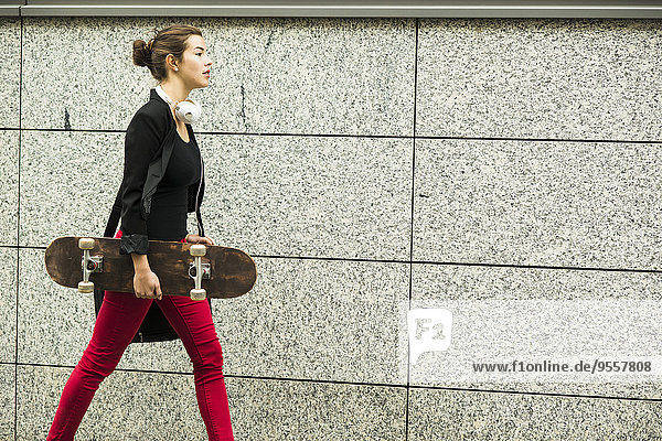 Junge Frau mit Kopfhörer und Skateboard läuft vor grauer Wand