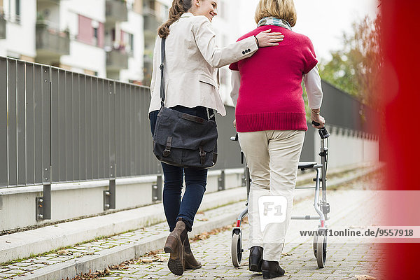 Adult granddaughter assisting her grandmother walking with wheeled walker  back view