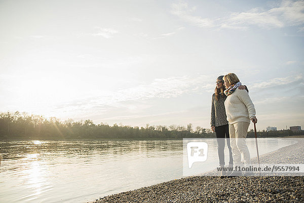 Junge Frau und Großmutter stehen zusammen am Wasser und beobachten den Sonnenuntergang.