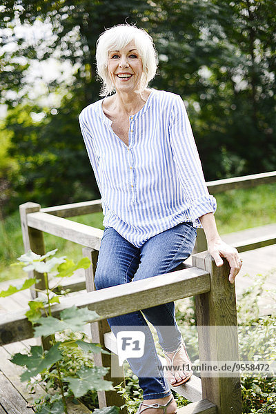Porträt der lächelnden weißhaarigen Seniorin auf Holzgeländer sitzend