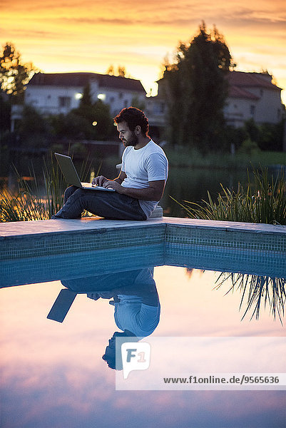 Man using laptop computer next to swimming pool as sun sets