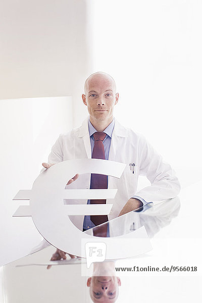 Porträt eines selbstbewussten Arztes mit Euro-Symbol am Schreibtisch in der Klinik