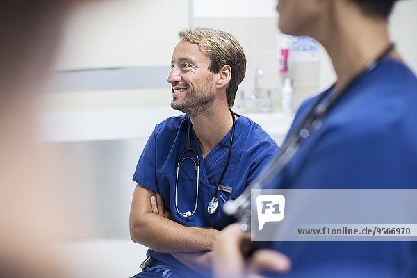 Mittlerer erwachsener Arzt mit verschränkten Armen sitzend und lächelnd in der Spitalabteilung