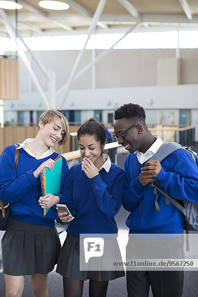 Drei glückliche Schüler in Schuluniformen mit Smartphone im Schulflur