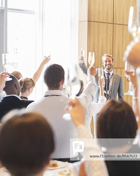 Porträt eines lächelnden Mannes  der vor Publikum im Konferenzraum steht und mit Champagnerflöte anstößt.