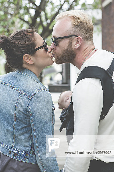View of couple in sunglasses kissing and holding baby in city streets
