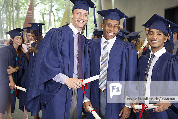 Portrait of three smiling male students in graduation gowns holding diplomas