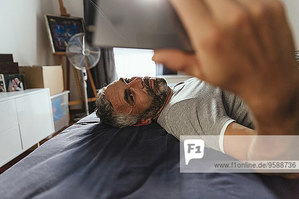 Ein ernsthaft aussehender Mann  der auf seinem Bett liegt und einen Selfie mit seinem Smartphone nimmt.