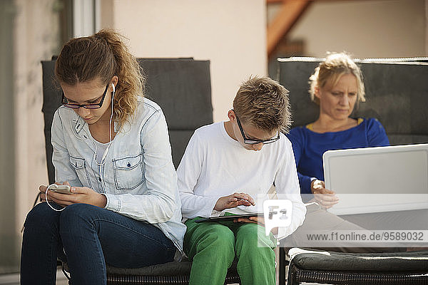 Mutter und ihre beiden Kinder entspannen sich mit digitalem Tablett  Smartphone und Laptop auf der Terrasse.