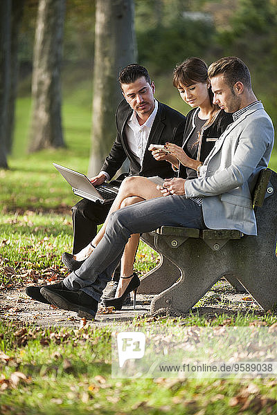 Drei Geschäftsleute sitzen auf einer Parkbank mit Laptop  Smartphone und digitalem Tablett.