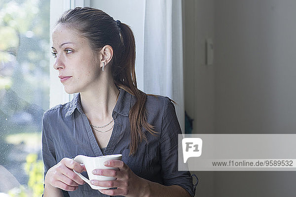 Junge Frau mit Tasse Kaffee durchs Fenster schauend