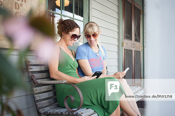 Zwei erwachsene Frauen,  die auf das Smartphone auf der Veranda schauen.