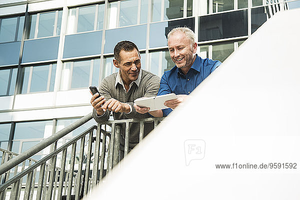 Two businessmen outside office building using cell phone and digital tablet