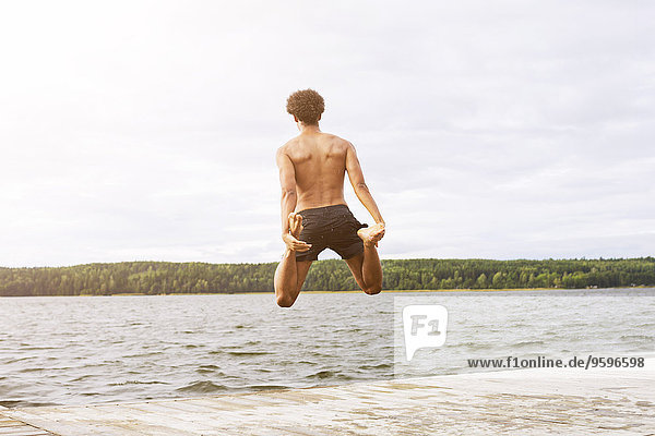 Full length rear view of man holding legs while jumping into lake