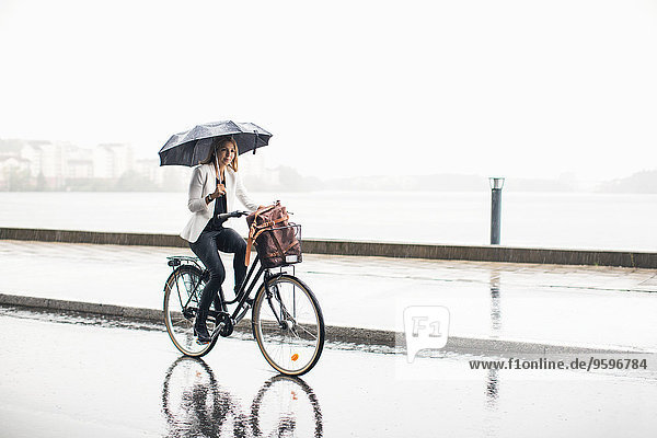 Volle Länge der Geschäftsfrau beim Radfahren auf der nassen Stadtstraße während der Regenzeit