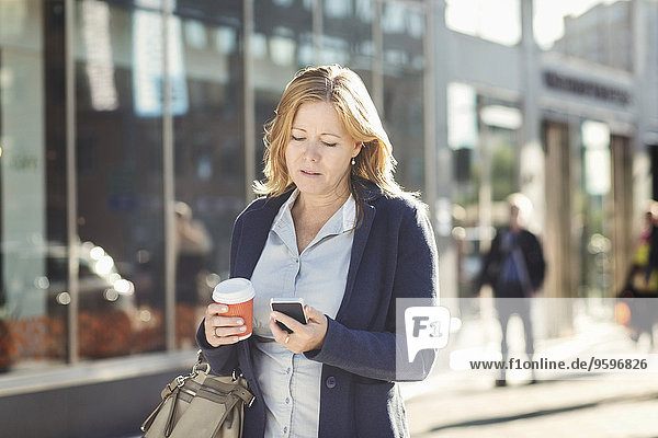 Geschäftsfrau mit Handy und Einweg-Kaffeetasse beim Gehen auf dem Bürgersteig