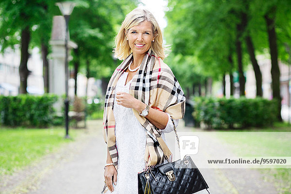 Portrait of smiling woman carrying purse while standing on footpath