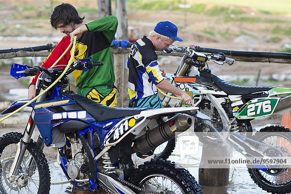 Zwei männliche Motocross-Teilnehmer beim Reinigen von Motorrädern mit Wasserschläuchen