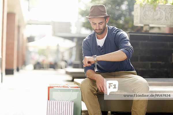 Mittlerer erwachsener Mann sitzt auf dem Sitz,  schaut auf die Uhr,  Einkaufstaschen neben ihm.