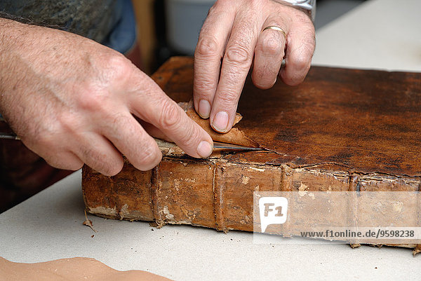 Nahaufnahme der Hände eines älteren männlichen Buchbinders  der Leder aus dem Buch entfernt.