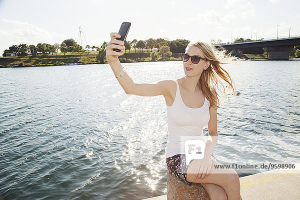 Young woman sitting at riverside taking smartphone selfie  Danube Island  Vienna  Austria