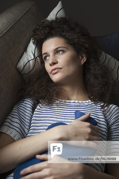Serious young woman lying on sofa with hot water bottle