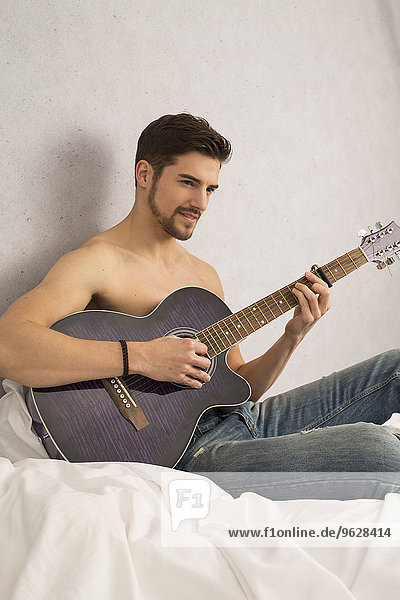 Shirtless man with guitar sitting on bed