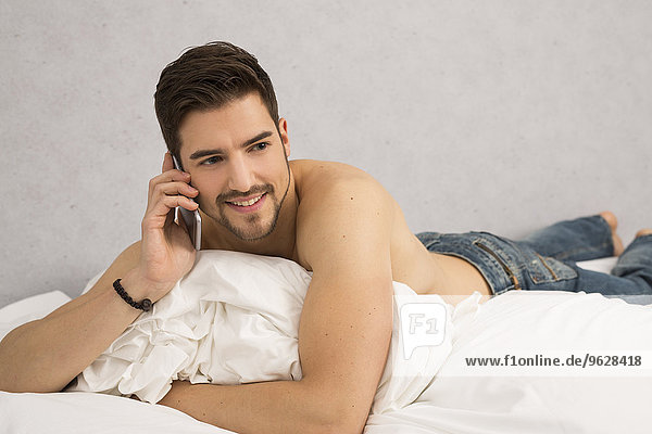 Shirtloser Mann auf dem Bett liegend telefoniert mit Smartphone