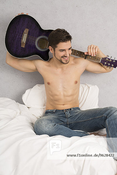 Shirtless man with guitar on his shoulders sitting on bed