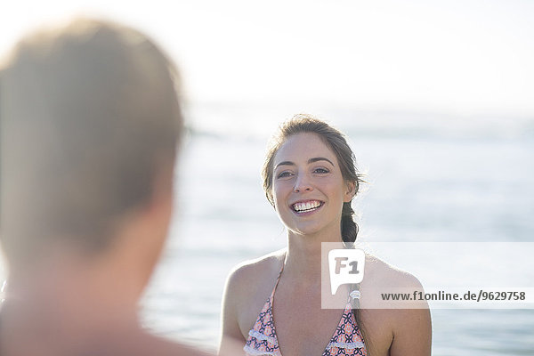 Lächelnde junge Frau am Strand mit Blick auf den Mann
