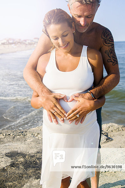 Pregnant mature woman and husband making heart shape on stomach at beach