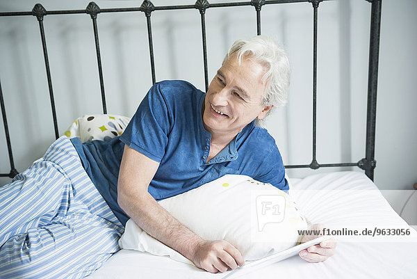 Mature man sitting with digital tablet in bed  smiling