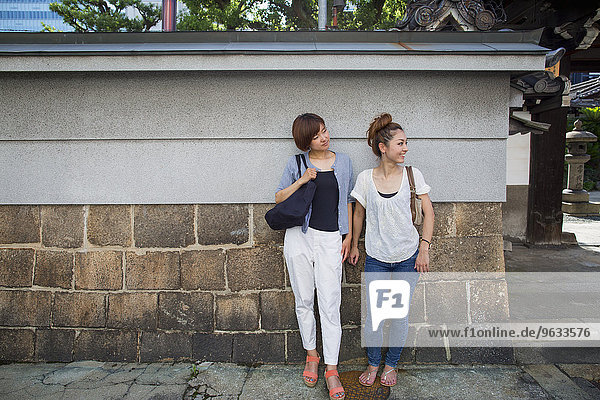 Two women standing outdoors  leaning against a wall.