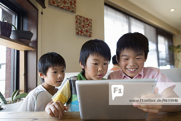 Three boys sitting at a table  looking at a digital tablet  smiling.