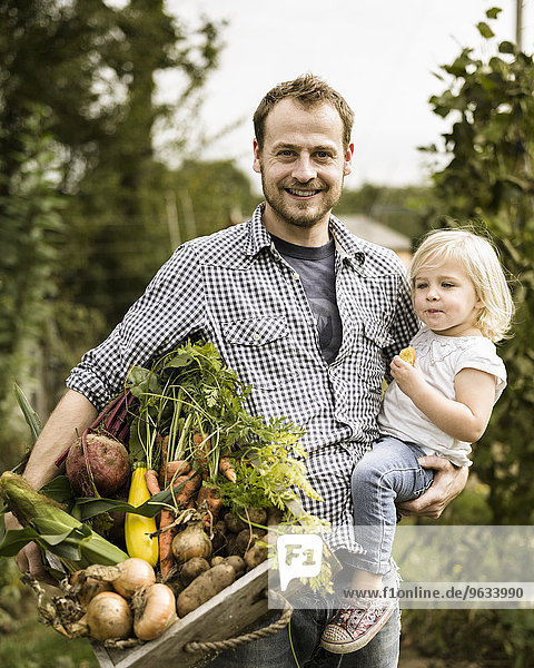 Man standing in his allotment with his daughter  smiling  holding a box full of freshly picked vegetables.