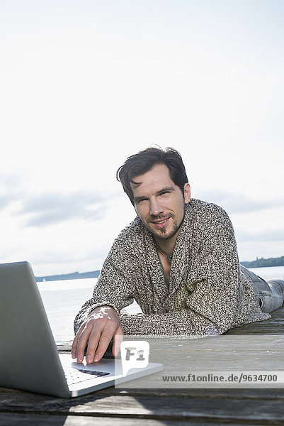 Portrait man lying on wooden jetty using laptop